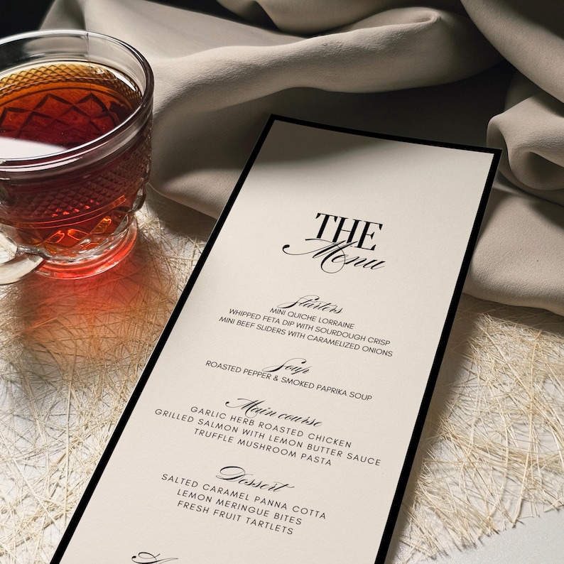 K&ouml;nnte beinhalten: Eine Speisekarte mit schwarzem Rand und dem Titel "THE Menu" in eleganter Schrift. Die Speisekarte listet Vorspeisen, Suppen, Hauptgerichte und Desserts auf. Links vom Men&uuml; steht eine Teetasse aus Glas mit einer dunklen Fl&uuml;ssigkeit.