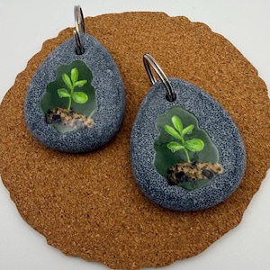 May include: Two gray stone-shaped keychains. Each keychain features a small, green plant design encased in clear resin. The keychains have silver-colored key rings attached at the top. The keychains are displayed on a brown, textured surface.