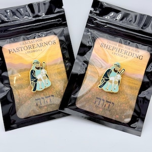 May include: Two enamel pins in clear packaging. Each pin features a shepherd holding a lamb, with text in English and Hebrew. The pins are on a card with a landscape background. The text on one pin reads "Thank you for your Shepherding".