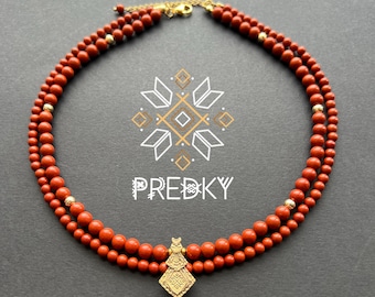 May include: A two-strand necklace featuring reddish-brown beads and gold-colored accents. A decorative pendant hangs at the center. The necklace is displayed on a dark gray surface. The word "PREDKY" is printed in white.