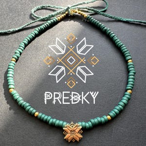 Op de afbeelding: Een groene en gouden kralenketting met een gouden sneeuwvlok hanger. De ketting ligt op een donkergrijze ondergrond met het woord "PREDKY" in wit gedrukt.