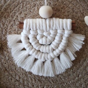 Può includere: Un arazzo da parete in macramè bianco con un design a semicerchio e frange. Una perlina di legno e uno spago sono attaccati in alto. L'immagine mostra anche perline di legno a forma di cuore e rotonde.