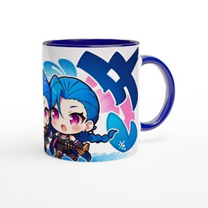 May include: A blue and white ceramic mug with a blue rim and handle. The mug features a cartoon illustration of two blue-haired characters with pink accents. The characters are on a white background with blue and pink graffiti-style markings.