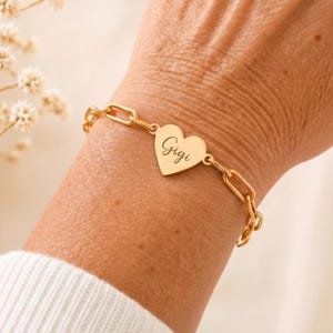 May include: A gold-colored bracelet with a heart-shaped pendant engraved with the name "Gigi". The bracelet features a chain link design. Additional images show the bracelet in silver, gold, and rose gold.