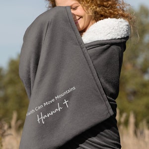 Könnte beinhalten: Eine dunkelgraue Decke mit weißem Sherpa-Futter. Auf der Decke steht der Text "Faith Can Move Mountains" und der Name "Hannah" mit einem Kreuzsymbol. Die Decke ist um eine Person gewickelt.