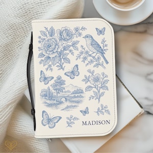 Puede incluir: Diario de color crema con un diseño floral y de mariposas azules. La portada presenta un pájaro posado en una rama, un paisaje y el nombre "MADISON". Incluye un asa negra.