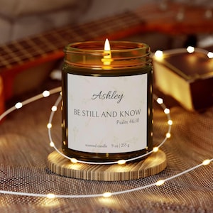 Puede incluir: Una vela de vidrio marrón con una etiqueta blanca que dice "Ashley Be Still and Know Psalm 46:10 scented candle 9 oz | 255 g". La vela está encendida y se encuentra sobre un posavasos de madera.