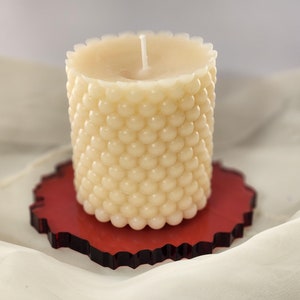 Könnte beinhalten: Eine cremefarbene, strukturierte Kerze mit runder Form und Docht, die auf einem roten, unregelmäßig geformten Untersetzer steht. Die Kerze hat eine blasenartige, strukturierte Oberfläche. Der Untersetzer ist tiefrot.