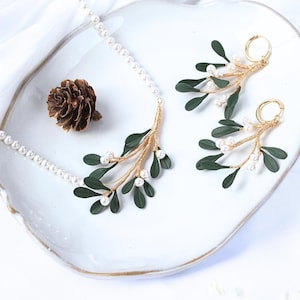 Könnte beinhalten: Ein Halsketten- und Ohrring-Set mit einem goldfarbenen Mistelzweig-Design, dunkelgrünen Blättern und kleinen weißen Perlen. Der Schmuck wird auf einem weißen Teller mit gewelltem Rand, vor weißem Hintergrund und einer Tannenzapfen präsentiert.