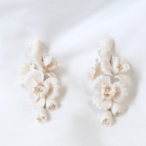 Op de afbeelding: Een paar witte keramische oorbellen in de vorm van delicate bloemclusters. De oorbellen zijn gemaakt van glad, mat wit keramiek en hebben ingewikkelde details.