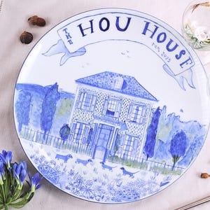 Könnte beinhalten: Weißer Keramikteller mit einer blauen Illustration eines Hauses, Hunden und floralen Elementen. Der Teller trägt den blauen Text "THE HOU HOUSE ren, 2023". Eine Gabel, ein Löffel und blaue Blumen sind um den Teller angeordnet.