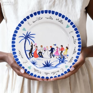 May include: A white ceramic plate with a blue scalloped border featuring a hand-painted illustration of a group of people dancing near a palm tree. The Italian text "È scritto nelle stelle" is written in black script around the inner rim.