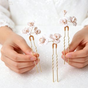 Peut inclure: Trois épingles à cheveux fleuries. Chaque épingle présente un bouquet de fleurs rose clair fixé à une base en fil d'or. Les épingles ont un design ondulé et sont tenues par une personne.