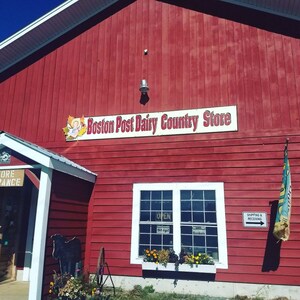 May include: A red wooden building with a white sign that reads "Boston Post Dairy Country Store". The building has a white window with a flower box and a green flag with a white design.