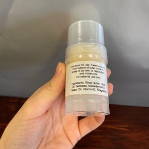 Puede incluir: Un tubo de pl&aacute;stico blanco con una etiqueta transparente que contiene una loci&oacute;n blanca. La etiqueta dice "Instrucciones de uso: Gire la loci&oacute;n hacia arriba desde la parte inferior del tubo. Aplique en &aacute;reas de piel seca para ayudar a suavizar e hidratar. Solo para uso externo. Ingredientes: Manteca de karit&eacute;, aceite de oliva, cera de abejas, aceite de macadamia, aceite de ricino, vitamina E, fragancia"