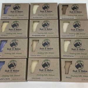 May include: Twelve brown cardboard boxes with clear plastic windows showcasing goat milk soap bars. Each box features a goat silhouette and the text "Back To Nature Goat Milk Soap" with the location "Enosburg Falls, Vermont" and the weight "Net Wt. 4.0 oz (113.4 g)". The soap bars are in various colors and scents, including Lavender Rose, Lily of the Valley, Sandalwood Vanilla, Lemongrass, Pomegranate Orange, Coconut Lime Verbena, Lilac, Cucumber Melon, Oatmeal Honey, Raspberry Vanilla, Baby Powder, and Almond.