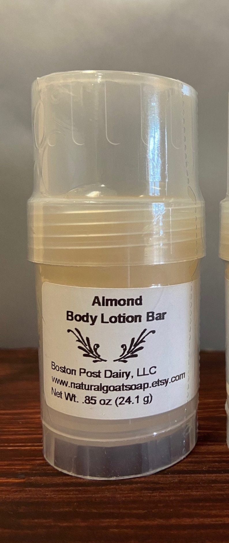 Puede incluir: Un recipiente de pl&aacute;stico blanco con una etiqueta transparente que dice "Almond Body Lotion Bar" y "Boston Post Dairy, LLC www.naturalgoatsoap.etsy.com Net Wt. .85 oz (24.1 g)".