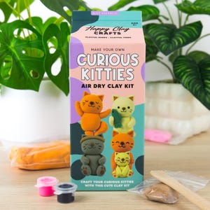 May include: A rectangular cardboard box for a Curious Kitties air dry clay kit. The box is light pink and teal with purple spots, featuring illustrations of four clay cats. Text includes Happy Clay Crafts, Make Your Own, and Curious Kitties.