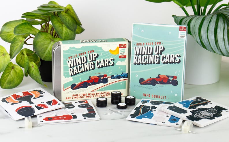 May include: A kit for building wind-up racing cars. The kit includes a tin box, an info booklet, car parts, and wheels. The box and booklet feature illustrations of red and blue race cars. The text on the box reads "Build Your Own Wind Up Racing Cars."