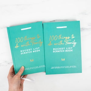 May include: Two teal "100 Things to Do with Family" bucket list scratch books. The covers have gold lettering that reads "100 things to do with family" and "Bucket List Scratch Book". The books feature interactive scratch pages.