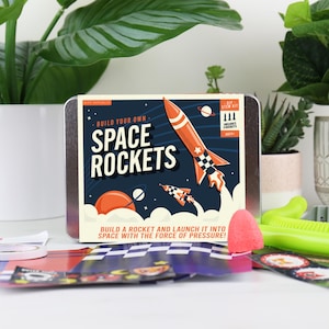 May include: A DIY STEM kit for building space rockets. The kit features a metal tin with the words "Build Your Own Space Rockets" and a graphic of rockets, planets, and stars. The kit also includes rocket parts and accessories.