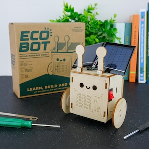 May include: A light-coloured wooden solar-powered robot kit with a smiling face, two circular antennae, and a black solar panel on top. The robot features a red power switch on the side and two wheels. The cardboard packaging is labelled ECO BOT.