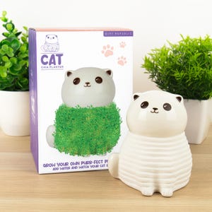 May include: A white ceramic cat-shaped chia planter with green 'fur' and a matching box. The box features a cat illustration and the text 'CAT CHIA PLANTER'. The planter is designed to grow chia seeds.