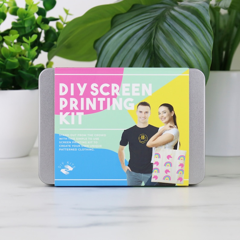Może przedstawiać: Srebrny pojemnik z kolorową etykietą, na kt&oacute;rej widnieje napis "DIY Screen Printing Kit". Etykieta przedstawia dwie osoby w koszulkach i torbę na ramię z wzorem tęczy. Na etykiecie widnieje r&oacute;wnież napis "Wyr&oacute;żnij się z tłumu dzięki temu łatwemu w użyciu zestawowi do sitodruku, aby stworzyć własne, niepowtarzalne ubrania z wzorami."