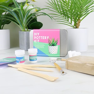 May include: DIY pottery kit with a white paint pot, sealant pot, clay, tools, and a tin box with a pink and blue design that says "DIY Pottery Kit".