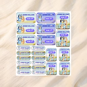 Puede incluir: Hoja de etiquetas personalizadas con personajes de dibujos animados. Cada etiqueta incluye el nombre "Joanna Williams" y "Grade 2nd". Las etiquetas tienen un esquema de color azul y morado, con personajes de dibujos animados.