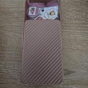 May include: A rectangular, light brown storage box with a textured, diagonal striped pattern on the front. The box is open, revealing small round stickers and a pink pouch. Some stickers have fruit and honey illustrations.