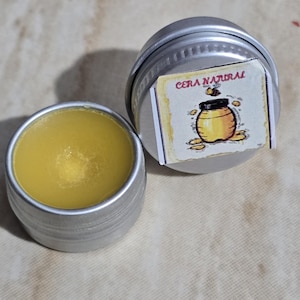 Puede incluir: Una pequeña lata de plata con cera amarilla en su interior. La tapa de la lata tiene una etiqueta que dice "CERA NATURAL" con una imagen de dibujos animados de un tarro de miel y una abeja.
