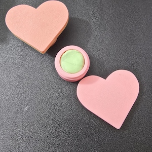 Op de afbeelding: Twee roze hartvormige make-upsponzen en een kleine roze container met een groen make-upproduct. De items zijn gerangschikt op een donkere ondergrond, wat een eenvoudige, strakke esthetiek creëert.