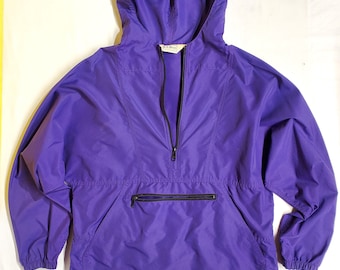 May include: A purple hooded anorak with a half-zip closure and a front pocket with a black zipper. The jacket has a drawstring hood and a small logo patch at the bottom. The anorak is made of a lightweight, water-resistant material.