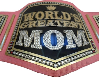 May include: A pink and gold championship belt with a black center plate that reads "World's Greatest Mom" in gold lettering with a crown above the text. The word "Mom" is adorned with rhinestones.