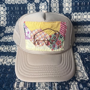 May include: A beige trucker hat with a patchwork design. The patchwork features yellow, red, and floral fabric pieces. The hat has a mesh back and a curved brim, offering a vintage aesthetic.