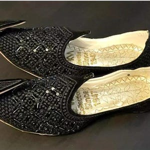 May include: A pair of black embroidered shoes with pointed toes and a decorative design. The shoes have a gold inscription on the inside.