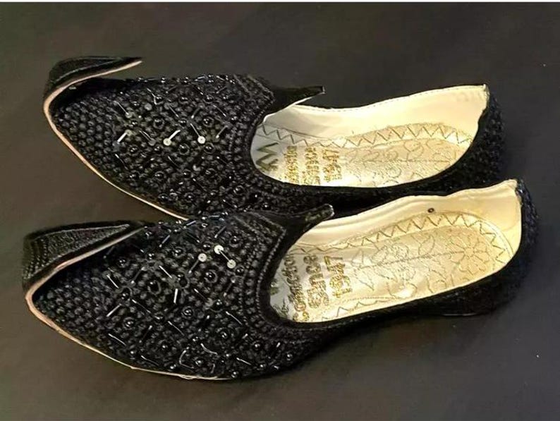May include: A pair of black embroidered shoes with pointed toes and a decorative design. The shoes have a gold inscription on the inside.