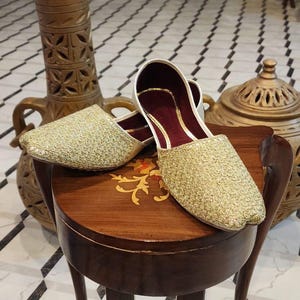 May include: A pair of gold embroidered slippers with maroon lining, sitting on a wooden table with intricate carvings. The slippers are on a white and black tiled floor.