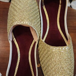 May include: A pair of gold embroidered flat shoes with maroon velvet lining. The shoes have a pointed toe and a slip-on design.