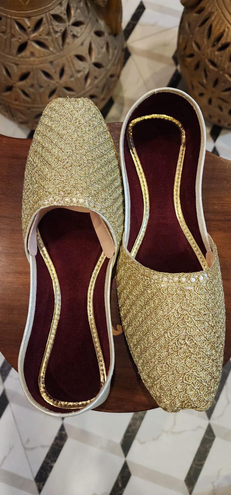 May include: A pair of gold embroidered flat shoes with maroon velvet lining. The shoes have a pointed toe and a slip-on design.