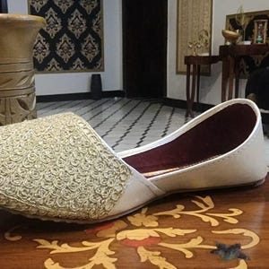 May include: A white embroidered flat shoe with gold thread. The shoe has a burgundy lining and is resting on a wooden surface.