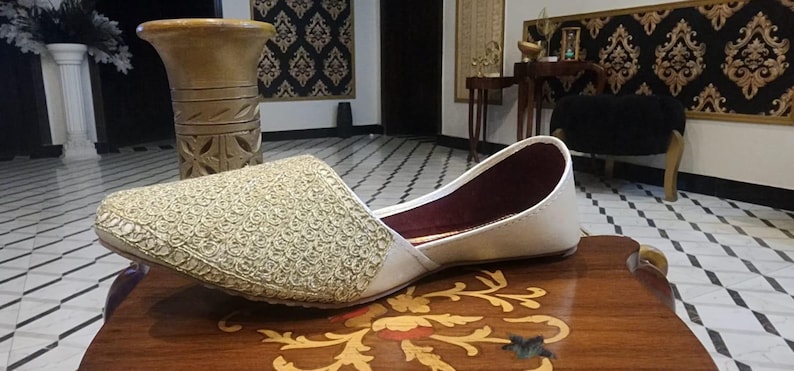 May include: A white embroidered flat shoe with gold thread. The shoe has a burgundy lining and is resting on a wooden surface.