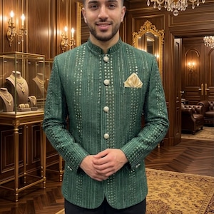 Könnte beinhalten: Eine grüne Jacke mit Stehkragen und aufwendigen silbernen Details. Die Jacke hat ein Einstecktuch und dekorative Knöpfe. Die Person trägt eine schwarze Hose. Der Hintergrund ist ein Raum mit goldenen Akzenten und einem Kronleuchter.