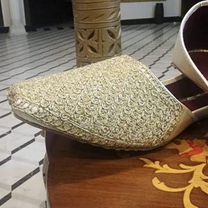 May include: A pair of white embroidered shoes with a gold pattern. The shoes are sitting on a wooden table with a gold inlay.