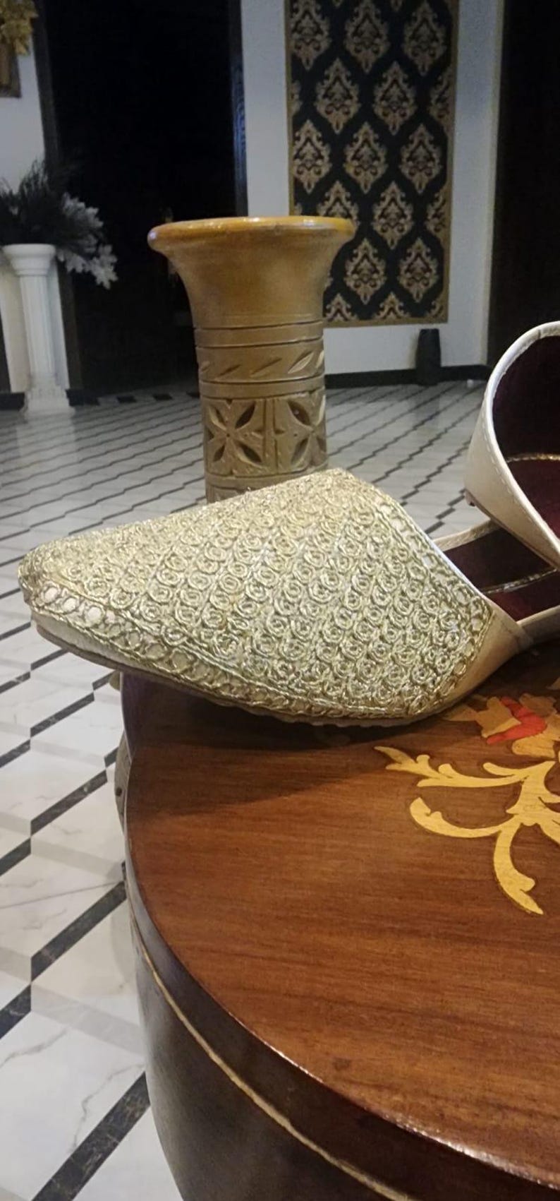May include: A pair of white embroidered shoes with a gold pattern. The shoes are sitting on a wooden table with a gold inlay.