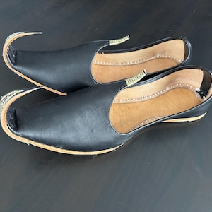 Puede incluir: Un par de zapatos de cuero negro con punta y un acento decorativo curvo. Los zapatos tienen una suela marrón claro y un interior beige. La parte superior del zapato es negra y el diseño es tradicional.