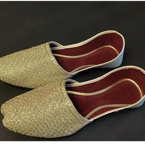 May include: A pair of gold embroidered flat shoes with maroon lining. The shoes have a pointed toe and a slip-on design.