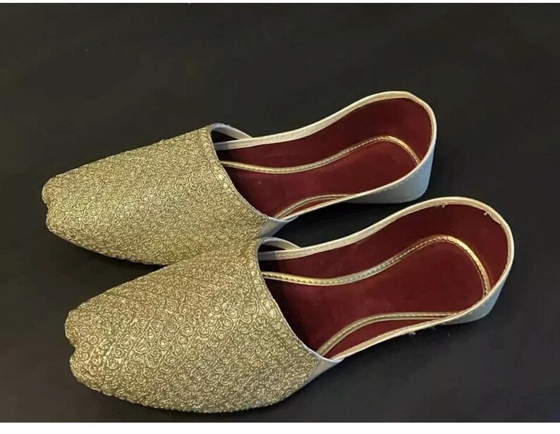 May include: A pair of gold embroidered flat shoes with maroon lining. The shoes have a pointed toe and a slip-on design.