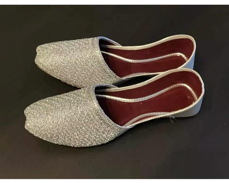 May include: A pair of silver sparkly flat shoes with a burgundy lining. The shoes have a pointed toe and a closed back.